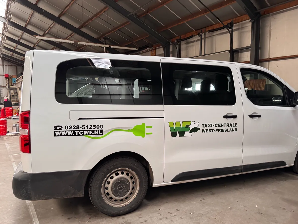 Witte bus in zijaanzicht met logo, telefoonnummer en groene stekker-lijn van Taxi-Centrale West-Friesland